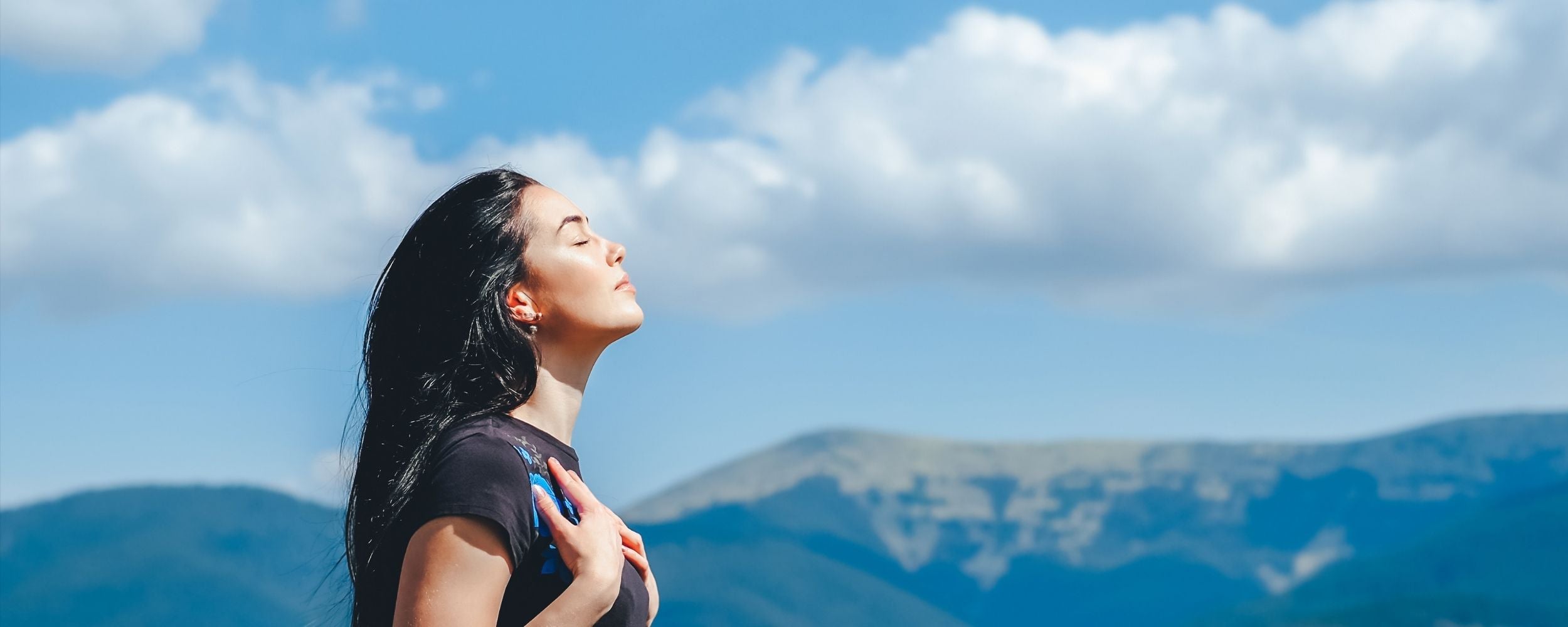 Woman with closed eyes against a blue sky with clouds and mountains in the background